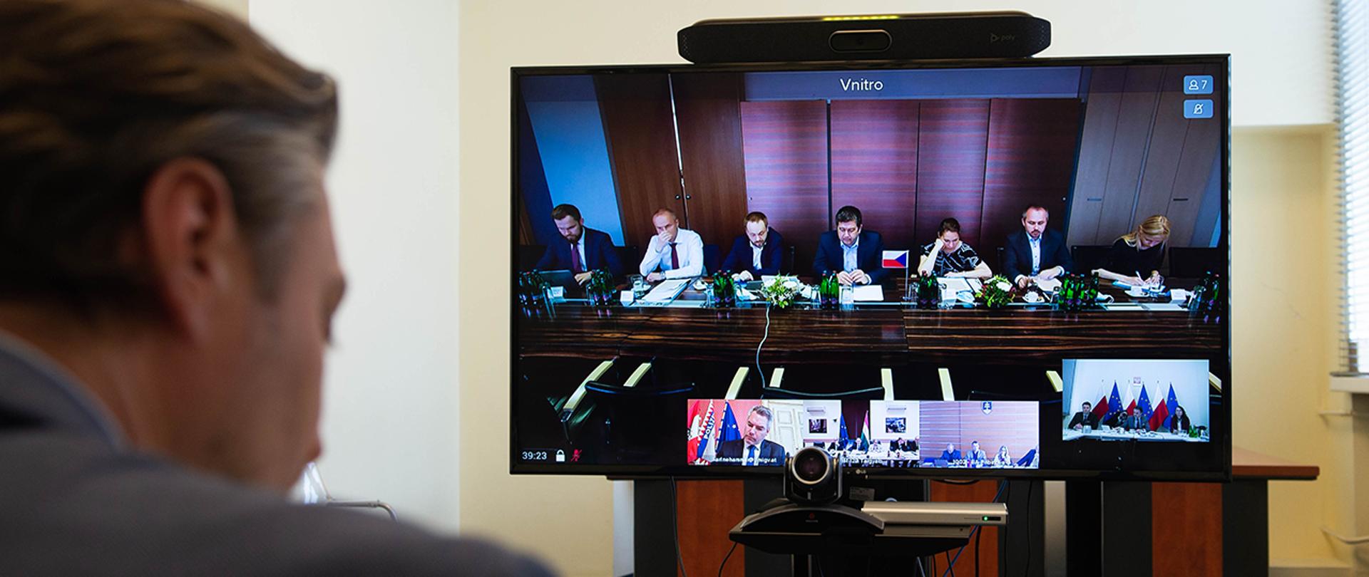 Ekran telewizora wyświetlający trwającą wideokonferencję z ministrami spraw wewnętrznych państw Grupy Wyszehradzkiej, ujęcie zza głowy wiceministra Bartosza Grodeckiego.