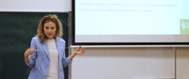Na tle szkolnej tablicy stoi młoda kobieta, która gestykuluje. Po prawej stronie widoczny slajd z prezentacją.