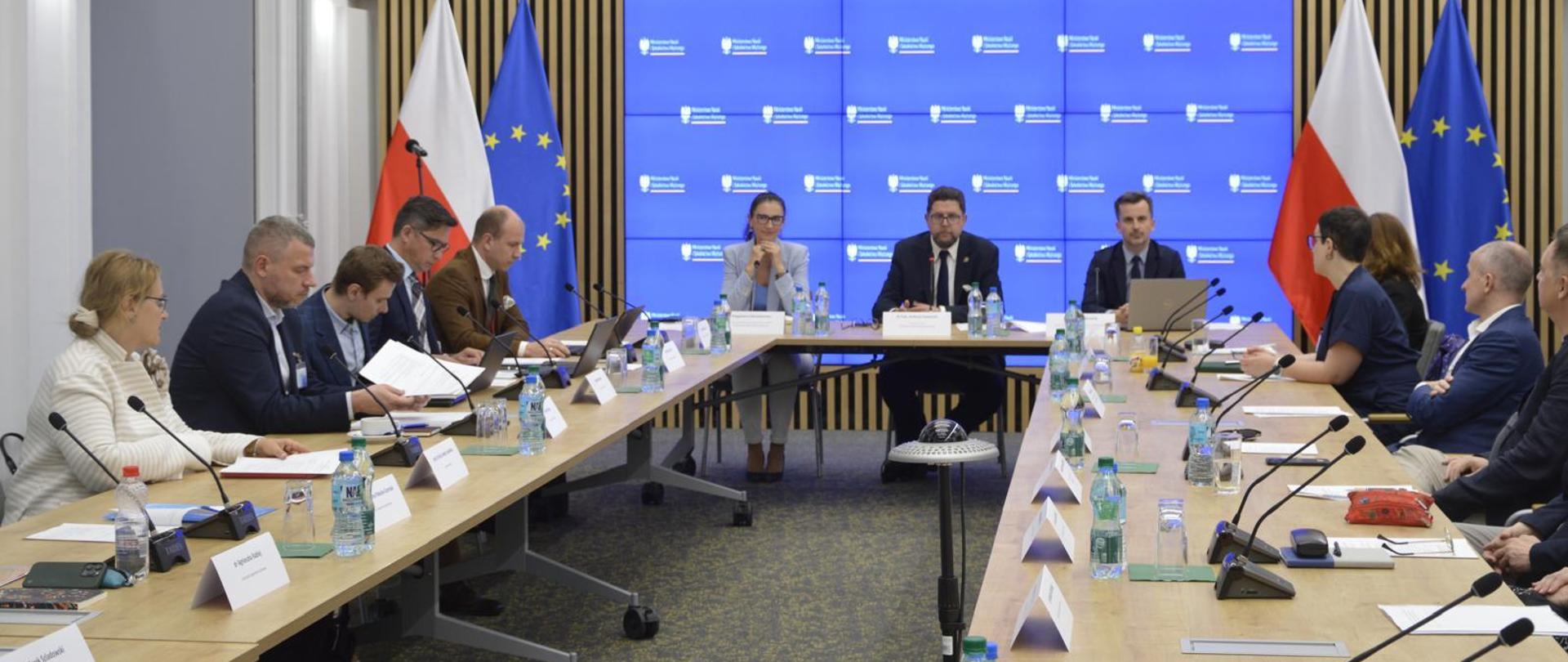 Za stołami ustawionymi w podkowę siedzą ludzie. Centralnie przy stole siedzi Wiceminister Andrzej Szeptycki