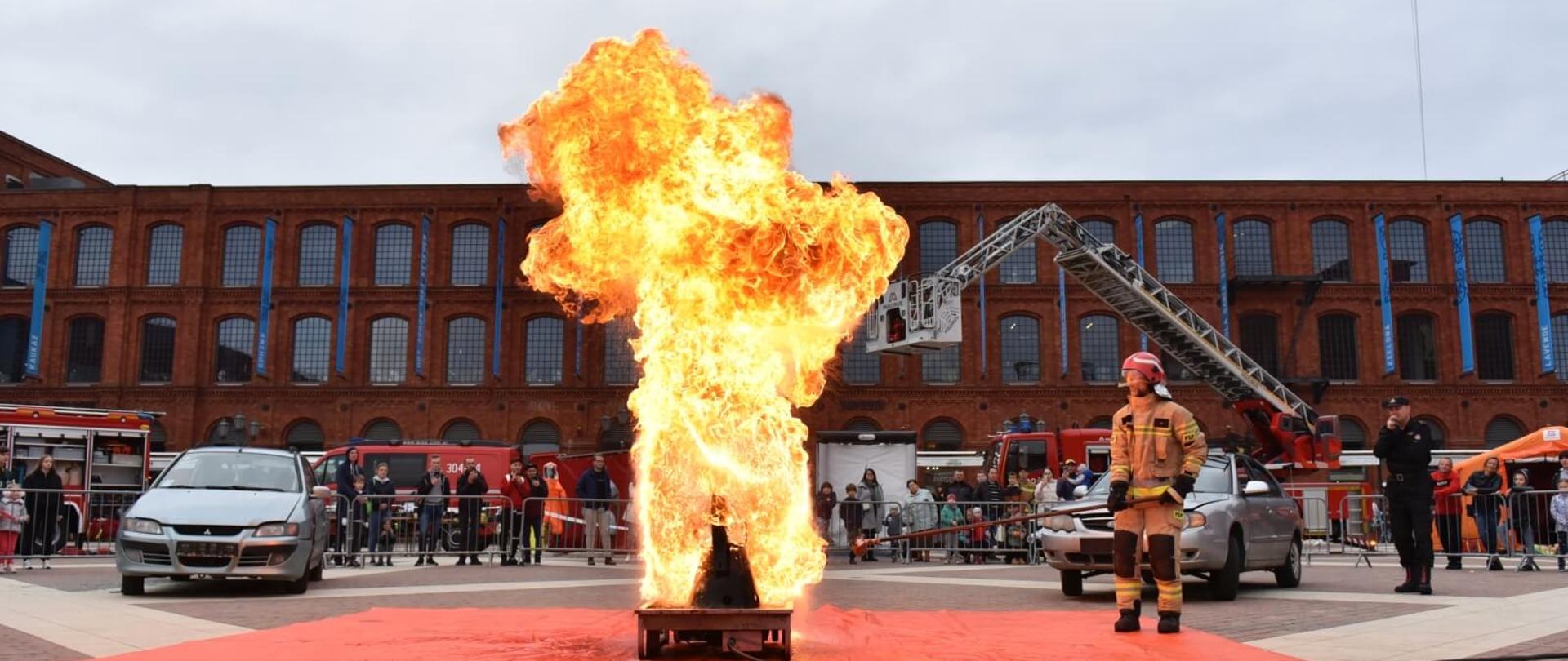 na głównym planie słup ognia nad metalowym urządzeniem obok strażak w mundurze bojowym w tle 2 wraki samochodów ludzie, samochody strażackie oraz budynek