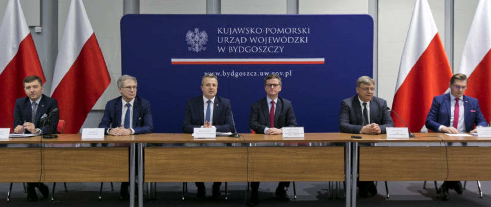 Na zdjęciu uczestnicy spotkania siedzą przy stole prezydialnym. W tle granatowa ścianka z napisem "Kujawsko-pomorski Urząd Wojewódzki w Bydgoszczy.