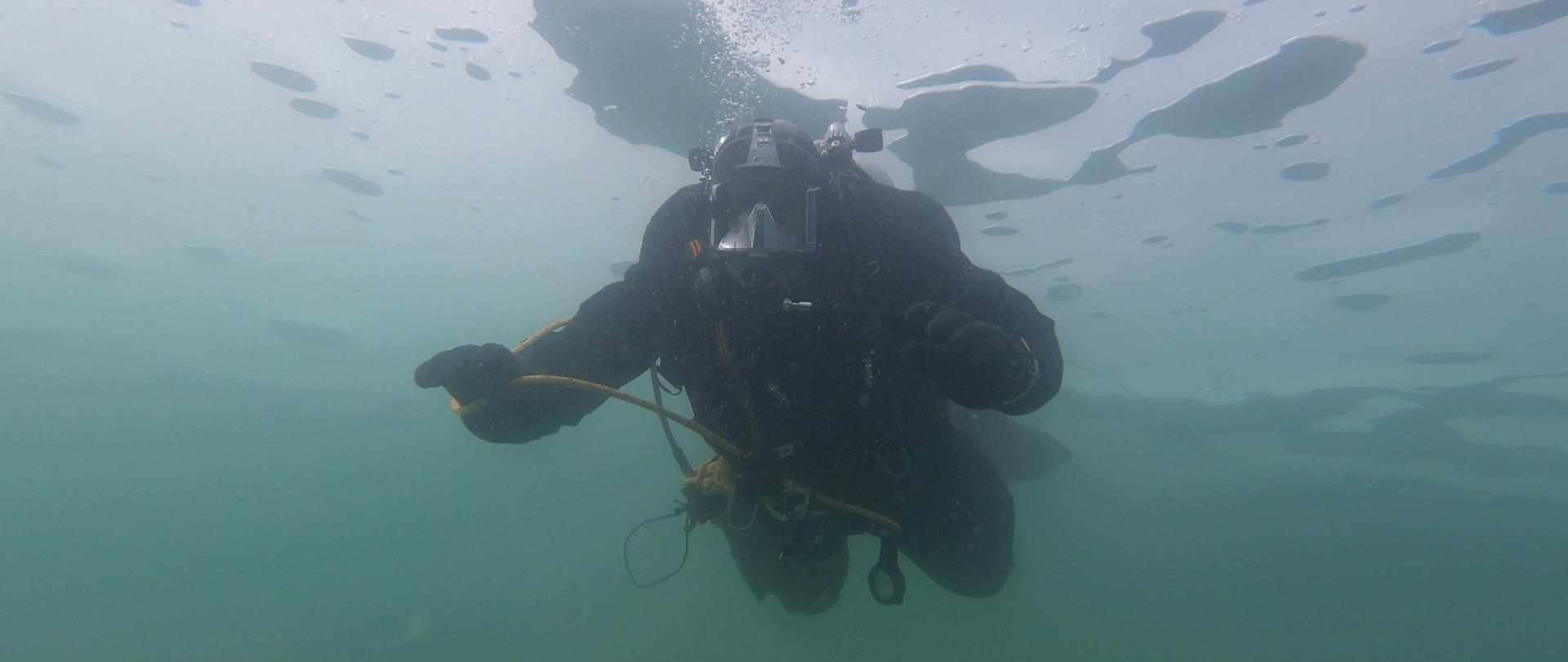 Zdjęcie zrobione pod wodą, przedstawia nurka w pełnym wyposażeniu, który nurkuje pod taflą lodu. Nad jego głową unoszą się pęcherzyki powietrza. 