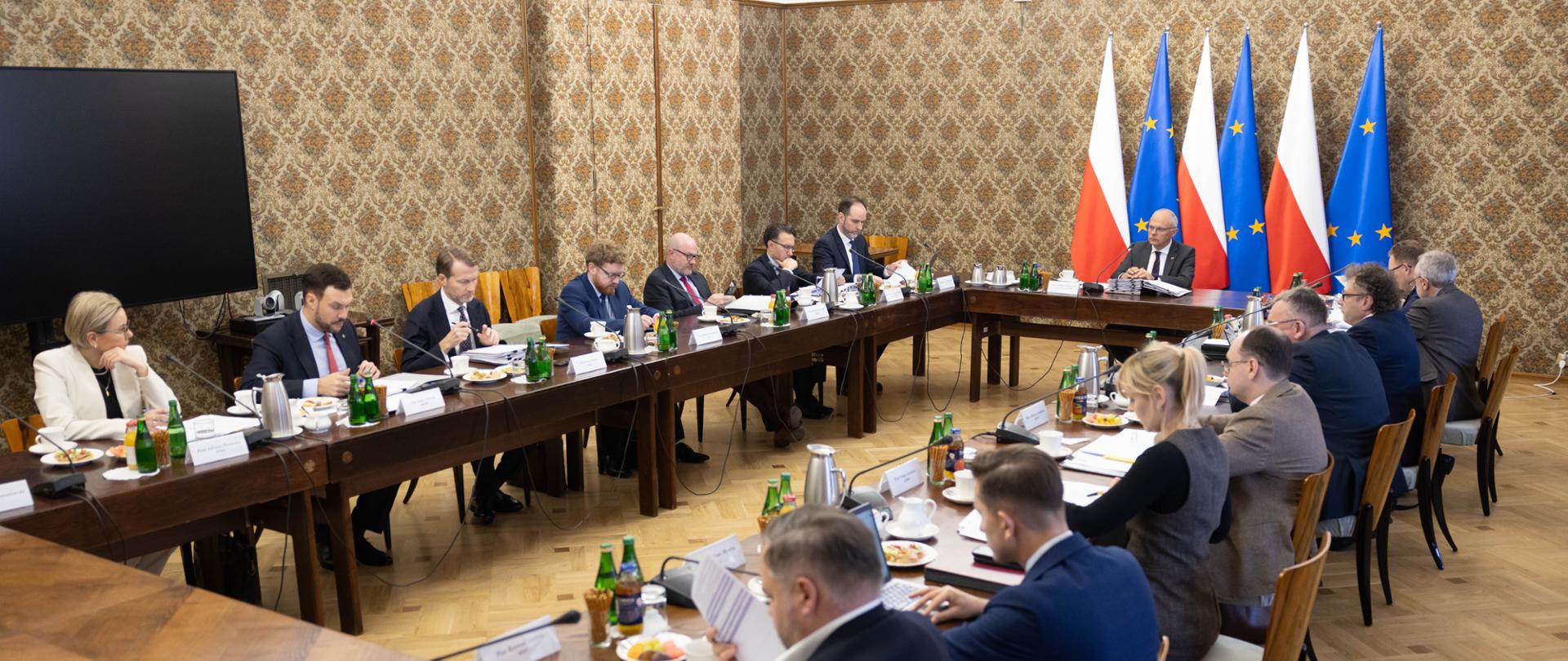 Posiedzenie Zespołu ds. Programowania Prac Rządu odbywające się przy dużym, drewnianym stole ustawionym w kształt litery U. Kilkanaście osób siedzi wokół stołów, przegląda dokumenty i prowadzi obrady. Na końcu sali znajdują się naprzemiennie ustawione flagi Polski i Unii Europejskiej.