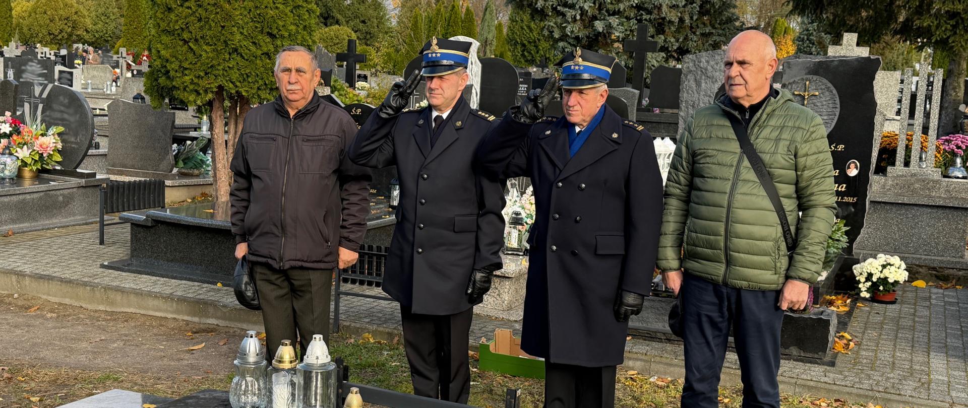 Czterech mężczyzn, w tym dwóch w mundurach strażackich, stoi przy nagrobku na cmentarzu. Jeden z mundurowych oddaje salut. Na grobie stoją zapalone znicze, a w tle widoczne są inne mogiły i drzewa.
