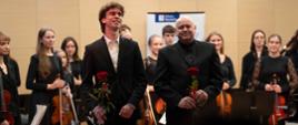 zdjęcie przedstawia ucznia szkoły oraz dyrygenta orkiestry trzymających kwiaty róży w dłoniach podczas oklasków po wykonaniu utworu, w tle orkiestra szkoły w pozycji stojącej oraz baner miasta Szczecin