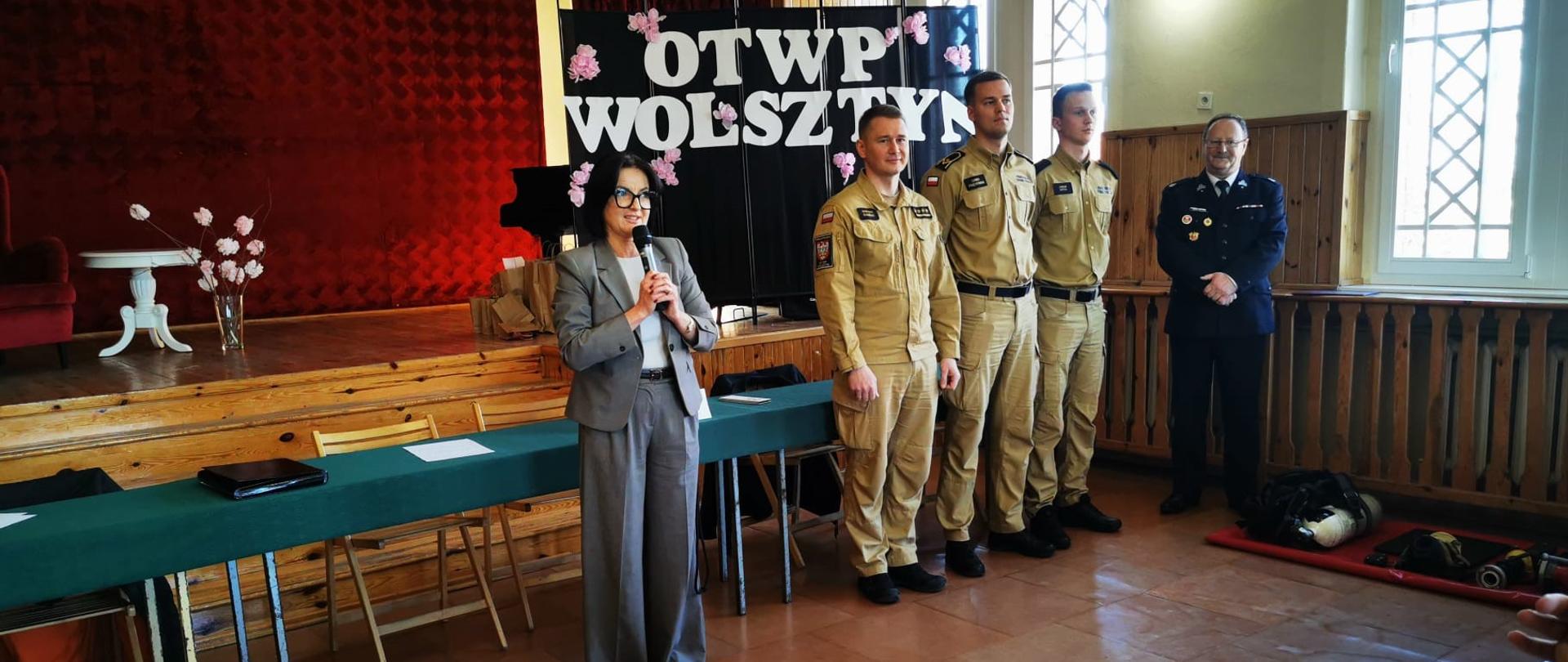 Na zdjęciu widać otwarcie konkursu Ogólnopolskiego Turnieju Wiedzy Pożarniczej. W centralnej części stoi kobieta przeprowadzająca oficjalne otwarcie konkursu. Po prawej stronie stoją funkcjonariusze Państwowej Straży Pożarnej na czele z zastępcą komendanta Powiatowego Państwowej Straży Pożarnej w Wolsztynie w piaskowych mundurach oraz druh Henryk Żok w mundurze galowym OSP. W tle widać scenę z czerwonym kurtynowym tłem, stolik z dekoracją (kwiaty w wazonie), a także okna wpuszczające światło dzienne oraz napis koloru białego ,,OTWP Wolsztyn”. Na podłodze, z prawej strony, leży sprzęt przypominający elementy wyposażenia strażackiego (np. aparat powietrzny).