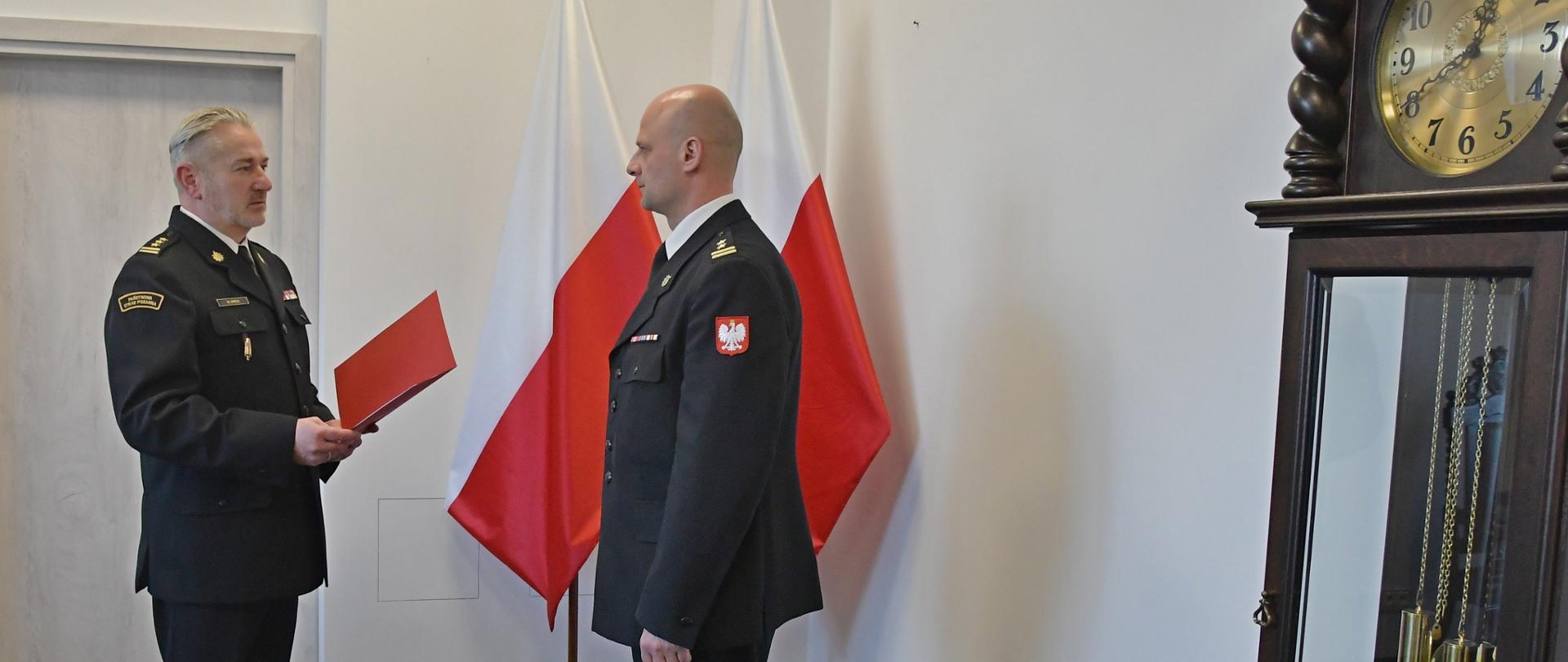 Pełniący obowiązki Pomorskiego Komendanta Wojewódzkiego Państwowej Straży Pożarnej starszy brygadier Marian Hinca wręcza decyzję o powierzeniu pełnienia obowiązków na stanowisku Komendanta Powiatowego Państwowej Straży Pożarnej w Lęborku brygadierowi Leszkowi Formela. Obok stoi duży zegar, w tle widoczne flagi państwowe.
