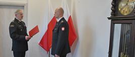 Pełniący obowiązki Pomorskiego Komendanta Wojewódzkiego Państwowej Straży Pożarnej starszy brygadier Marian Hinca wręcza decyzję o powierzeniu pełnienia obowiązków na stanowisku Komendanta Powiatowego Państwowej Straży Pożarnej w Lęborku brygadierowi Leszkowi Formela. Obok stoi duży zegar, w tle widoczne flagi państwowe.