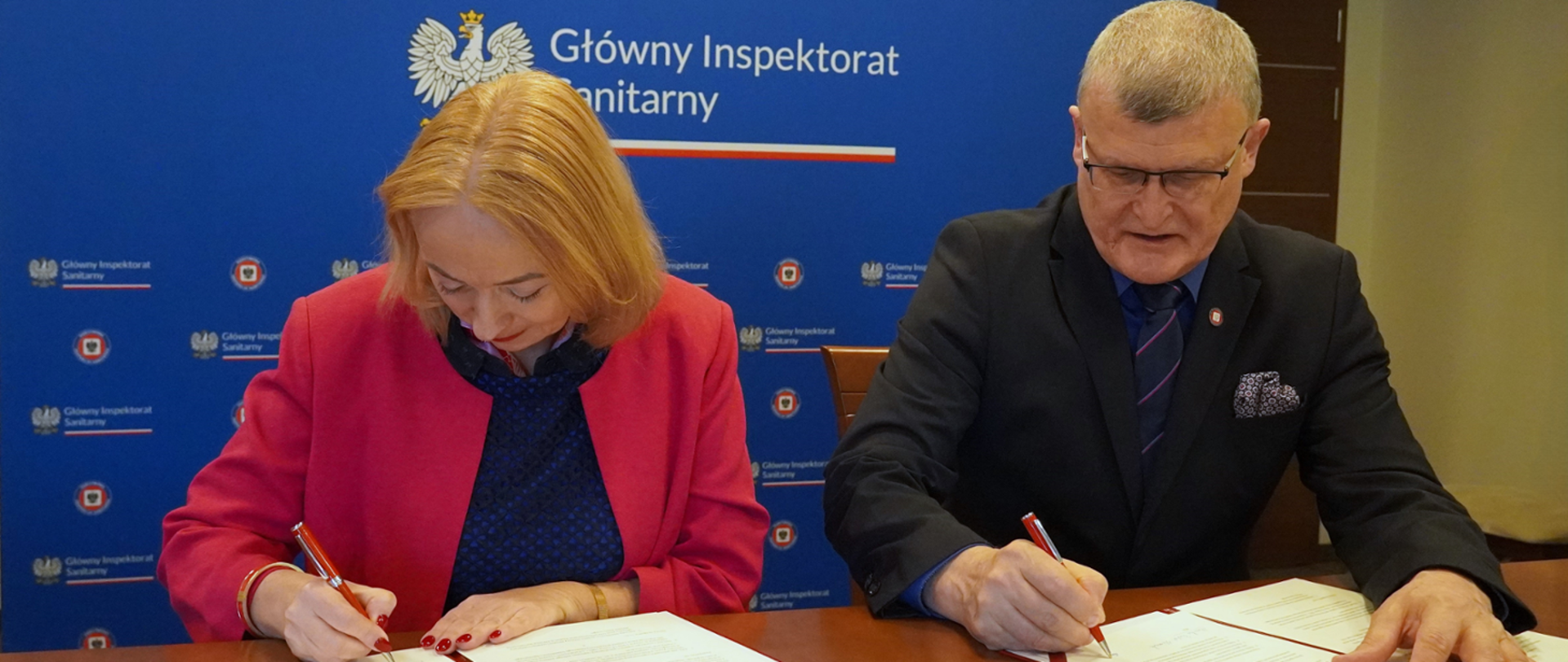 Na zdjęciu widzimy dwie osoby siedzące przy stole i podpisujące dokumenty. Kobieta po lewej stronie ma blond włosy i jest ubrana w granatową bluzkę oraz różową marynarkę. Mężczyzna po prawej stronie nosi ciemny garnitur, granatową koszulę i krawat w paski.
Oboje trzymają czerwone długopisy i koncentrują się na podpisywaniu dokumentów. W tle znajduje się niebieska tablica z logo i napisem „Główny Inspektorat Sanitarny”, co sugeruje, że wydarzenie ma oficjalny charakter i dotyczy kwestii związanych z ochroną zdrowia publicznego.
Zdjęcie przedstawia formalne spotkanie, które może dotyczyć współpracy między instytucjami zajmującymi się zdrowiem i bezpieczeństwem sanitarnym.