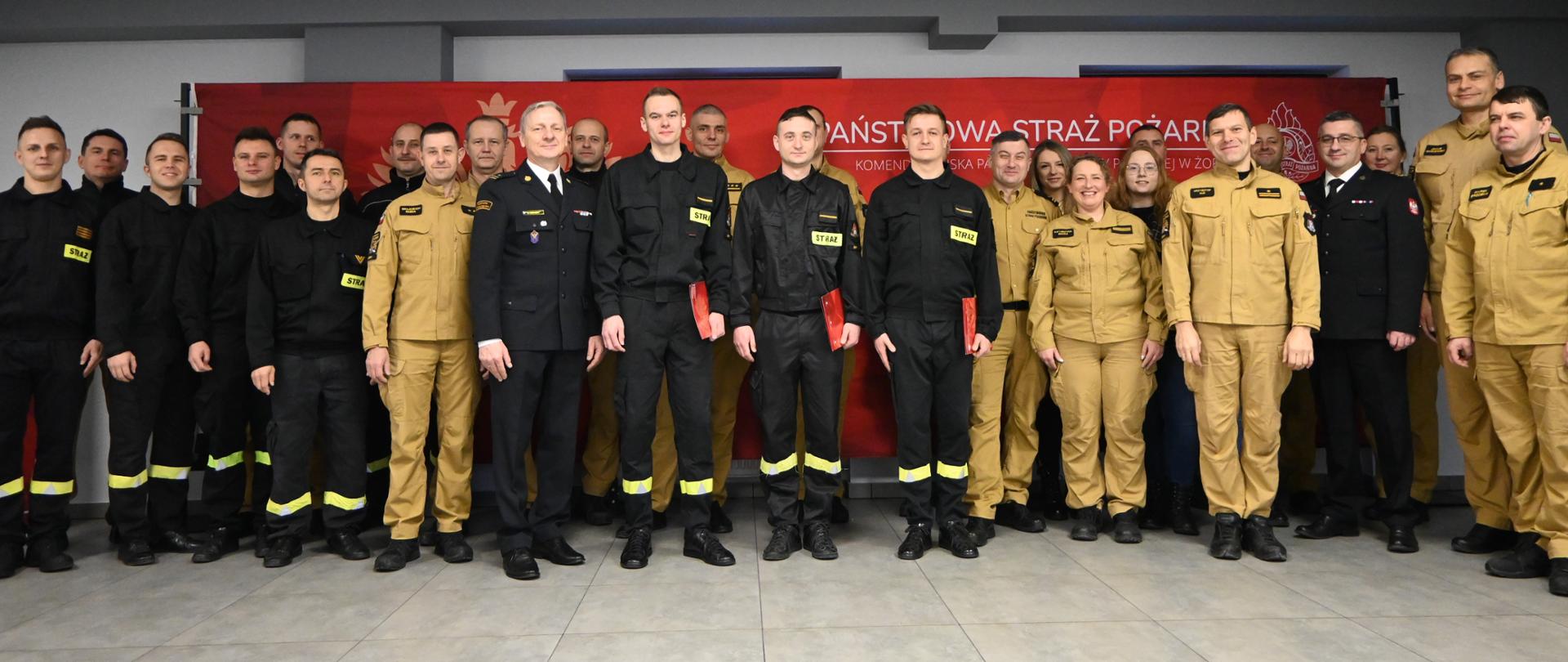 Pamiątkowe zdjęcie grupowe strażaków w mundurach służbowych i galowych, pozujących na tle czerwonego baneru z napisem „Państwowa Straż Pożarna”.