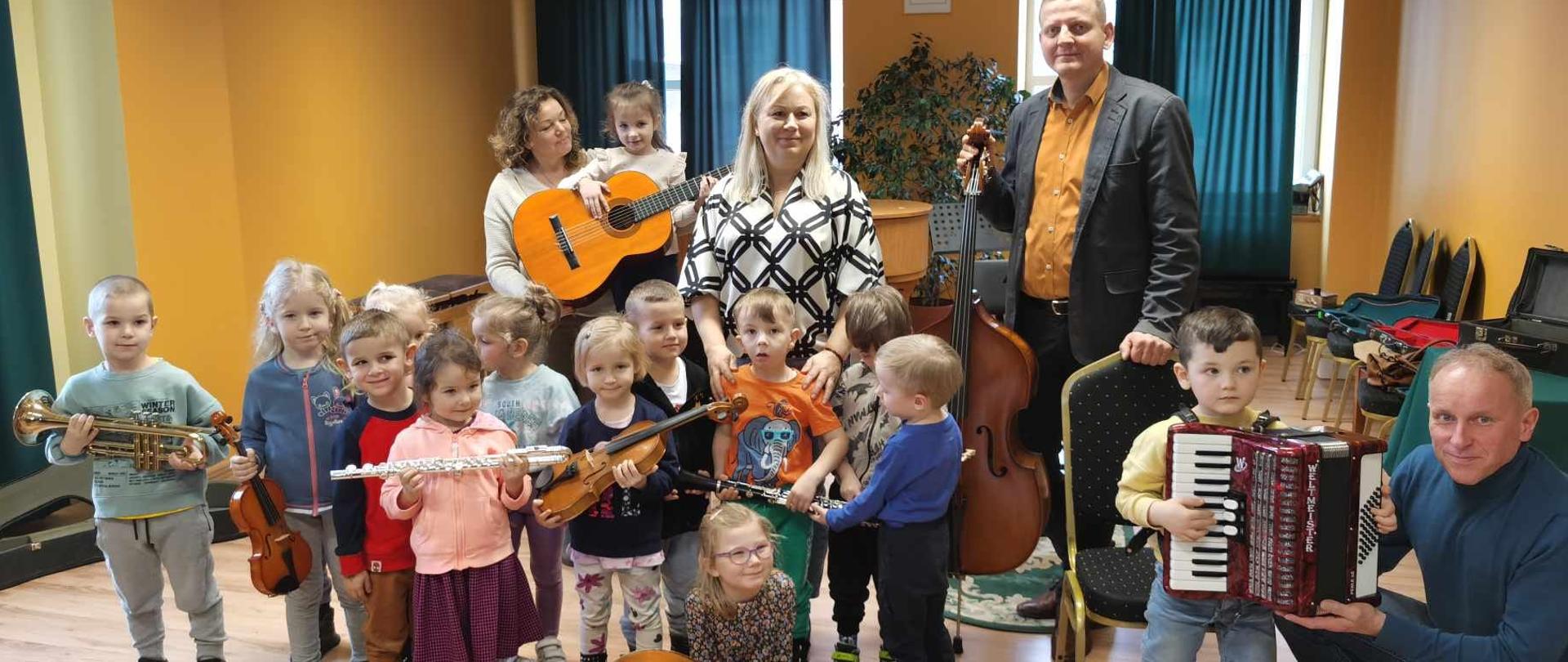 Zdjęcie grupowe. Grupa przedszkolaków stoi na środku sali trzymają w rękach instrumenty takie jak: trąbka, flet poprzeczny, skrzypce, klarnet. Chłopczyk po prawej stronie ma założony akordeon, który pomaga mu utrzymać nauczyciel kucający obok niego. Z tle za przedszkolakami stoi trzech nauczycieli. 