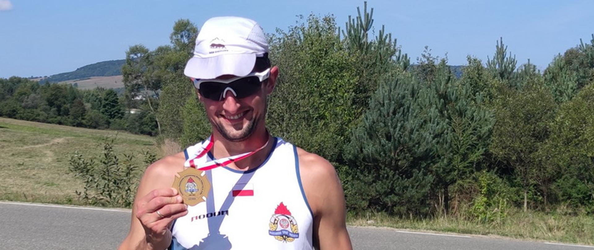 Mężczyzna w stroju sportowym z białą czapeczką na głowie i ciemnych okularach stoi trzymając w prawej ręce medal, w tle widoczny zarys gór