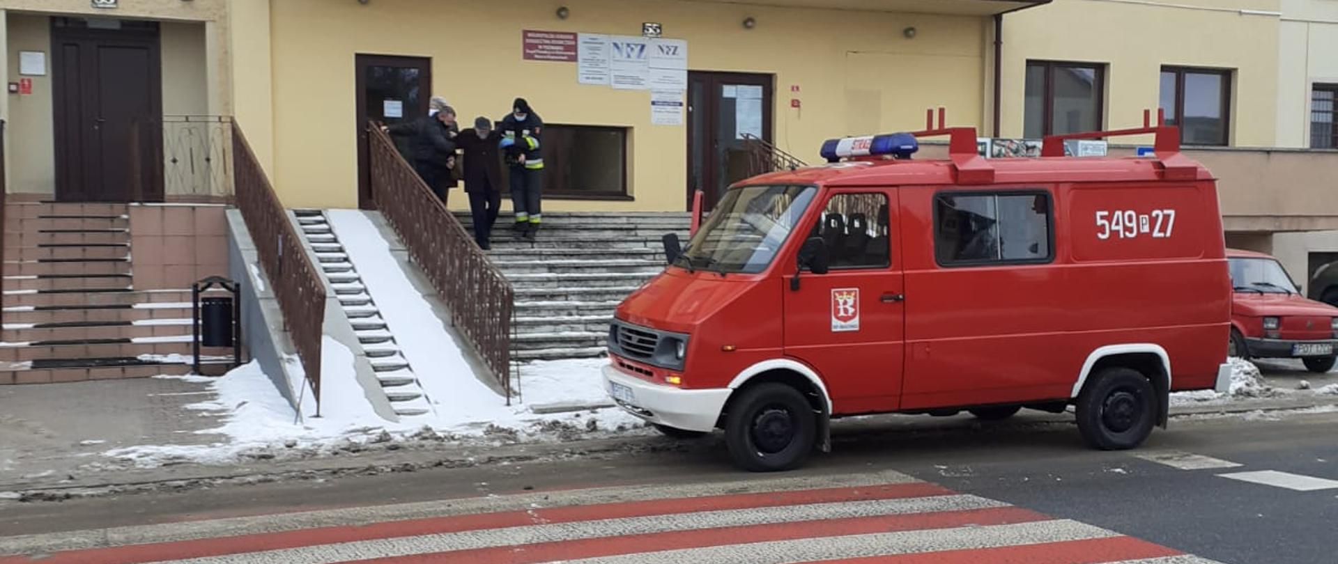 Zdjęcie przedstawia pomoc w transporcie seniora z punktu szczepień do pojazdu. Na zdjęciu widzimy strażaków ochotników, którzy pomagają seniorowi w zejściu po schodach, na pierwszym planie widoczny lekki samochód pożarniczy. Zdjęcie wykonano w porze dziennej na ulicy.