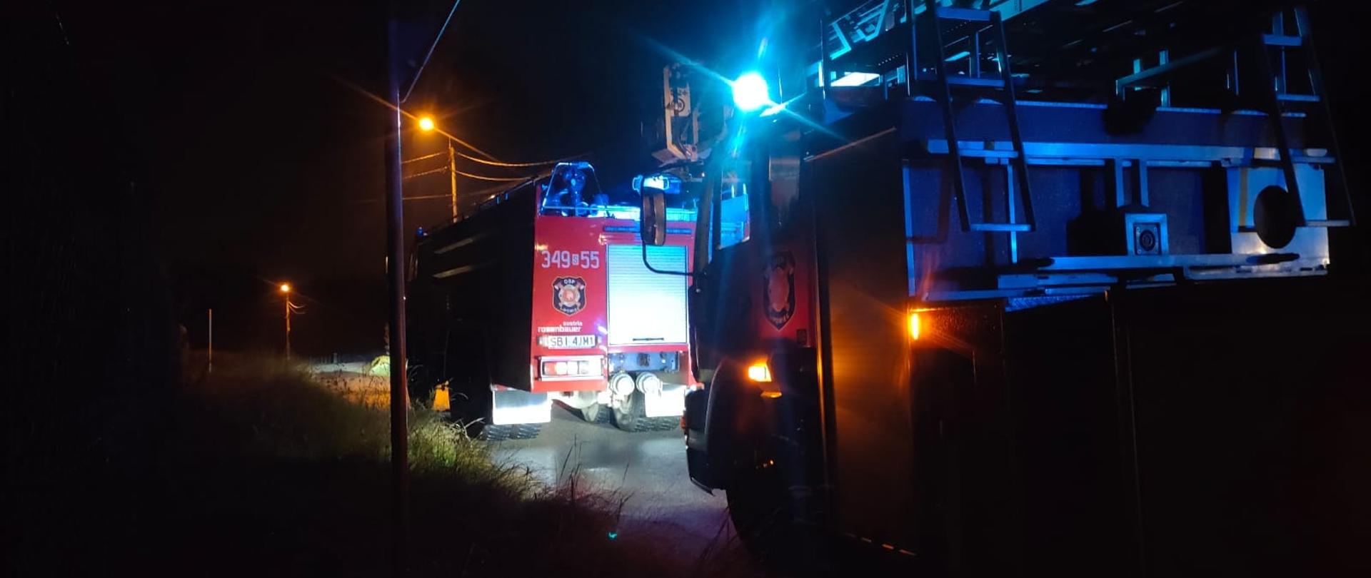 Dwa zastępy straży pożarnej stojące na drodze w nocy. 