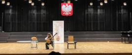 Sala koncertowa Zespołu Szkół Muzycznych w Kielcach, ogromna scena. Na środku sceny występuje uczennica naszej szkoły Amelia Lewkowicz podczas X Ogólnopolskiego Festiwalu i Konkursu "Gitara Viva" w dniu 16 marca 2023 r. Na środku ściany wisi duże godło Polski, na suficie wisi mnóstwo żyrandoli. Uczennica siedzi na krześle po środku sceny, podczas występu, po jej prawej i lewej stronie stoją dwa krzesła, za nią baner z napisem "Gitara Viva!"