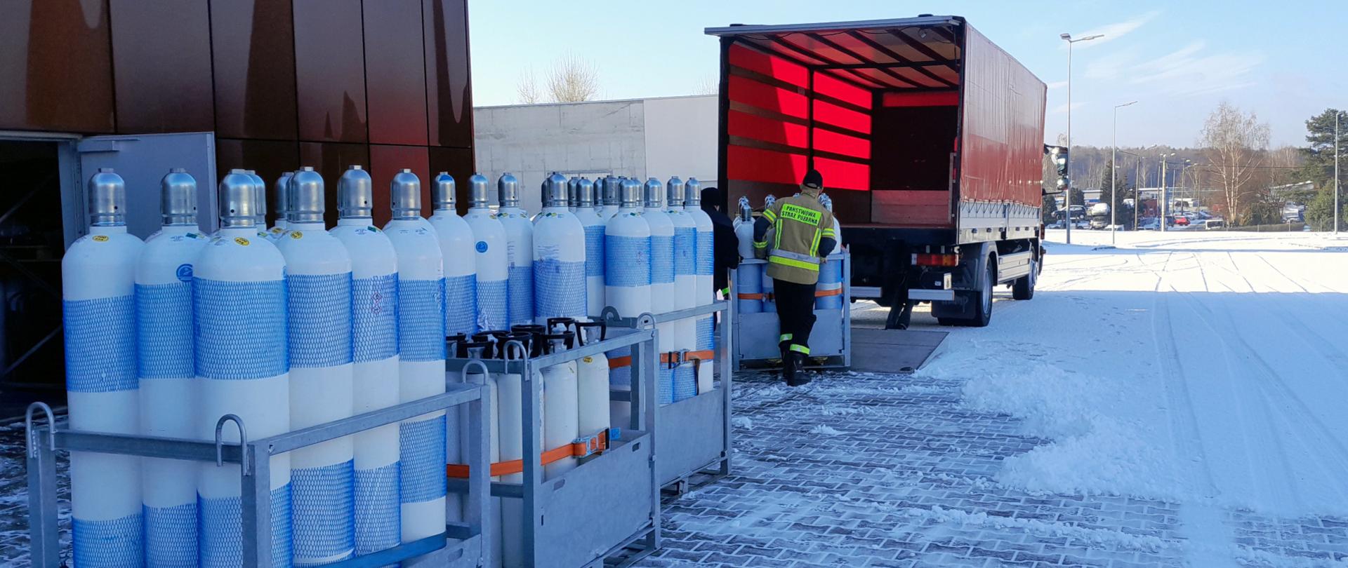 Strażak ubrany w kurtkę w kolorze żółtym z napisem na plecach państwowa straż pożarna ładuje do samochodu dostawczego butle z tlenem medycznym, samochód zaparkowany przed magazynem komendy Państwowej Straży Pożarnej w Mysłowicach. Za nim w rzędach ustawione są butle z tlenem.