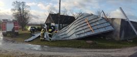 Zdjęcie przedstawia budynek gospodarczy z całkowicie zerwanym dachem, oraz strażaków z drabinami pożarniczymi w trakcie zabezpieczania dachu przed opadami.