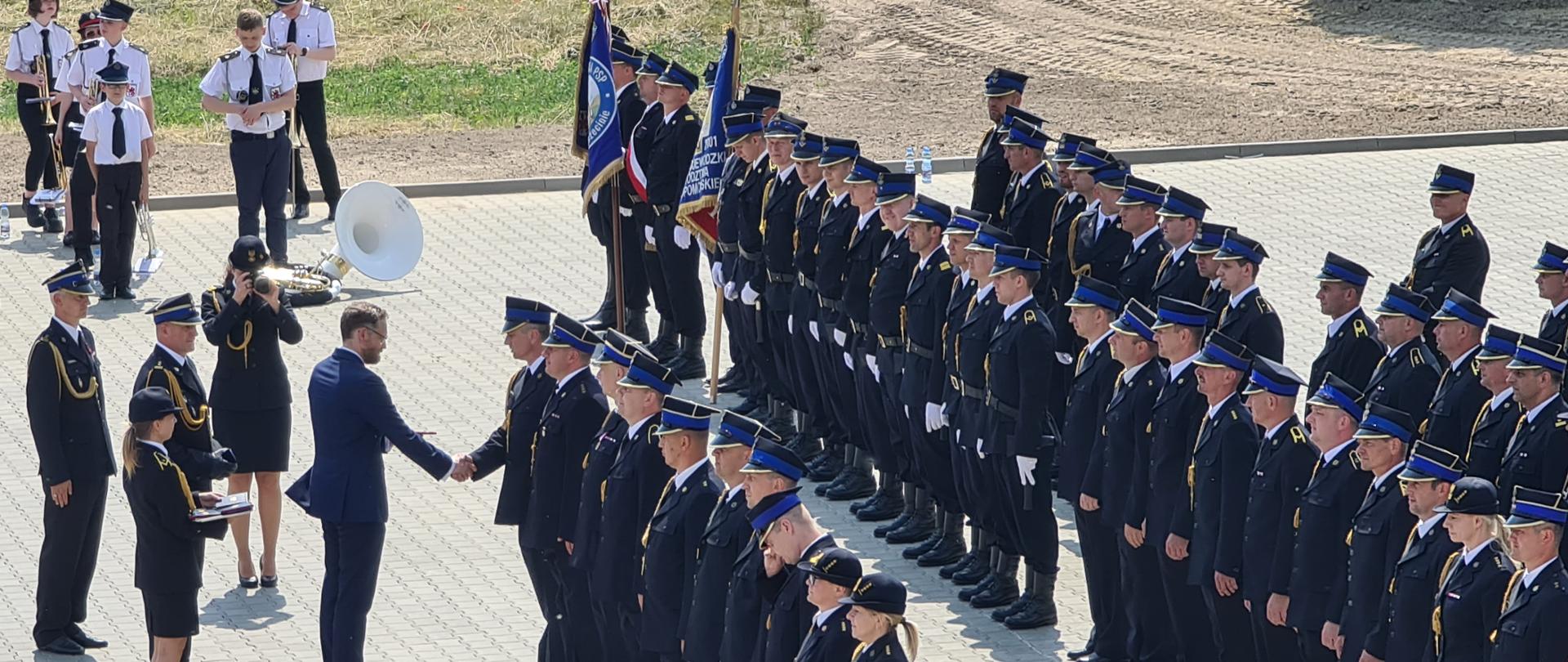 Zdjęcie wykonane z góry, z widokiem na plac na którym znajdują się po prawej stronie odznaczeni lub awansowani funkcjonariusze. W górnej części zdjęcia widoczna jest orkiestra OSP, w centralnej części zdjęcia znajdują się funkcjonariusze obecnie odznaczani. 