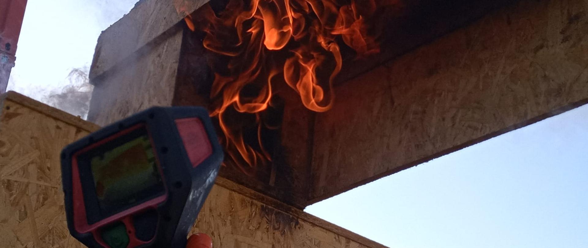 Alt: Zbliżenie na rękę w rękawicy trzymającą kamerę termowizyjną skierowaną na drewnianą konstrukcję, z której wydobywa się dym. Na ekranie kamery widoczny obraz termiczny z oznaczeniem temperatur.
