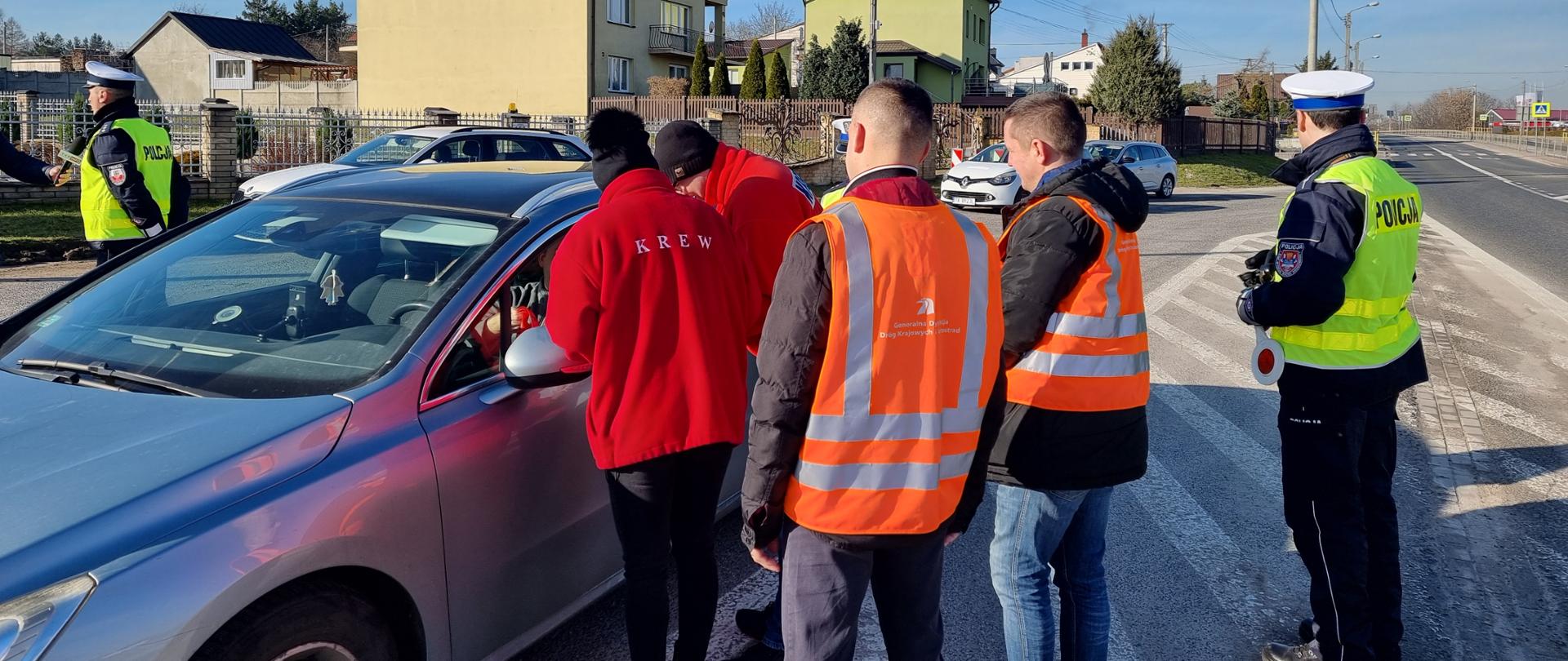 Drogocenna krew - samochód na parkingu, policjant z lizakiem w kamizelce odblaskowej, dwóch drogowców w kamizelkach pomarańczowych i dwoje pracowników stacji krwiodawstwa w czerwonych kurtkach z napisem "krew". Grupa rozmawia z kierowcą samochodu