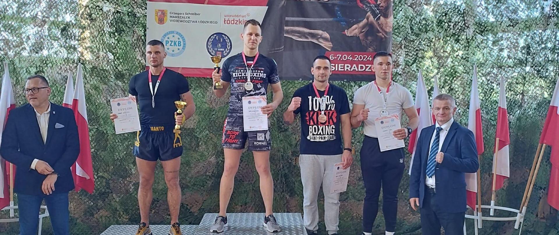 Na podium stoi czterech zawodników w sportowych strojach, po obu bokach podium stoją ubrani w garnitury mężczyźni.