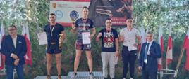 Na podium stoi czterech zawodników w sportowych strojach, po obu bokach podium stoją ubrani w garnitury mężczyźni.
