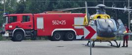 Zdjęcie przedstawia śmigłowiec LPR na rondzie. Śmigłowiec koloru żółtego, w tle samochód pożarniczy koloru czerwonego.