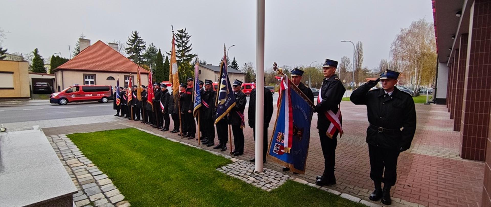 Strażacy stoją w szeregu przed pomnikiem strażaka.