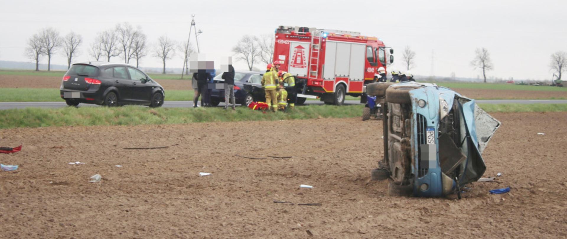 Samochód leży na boku na polu. W tle stoi samochód pożarniczy.