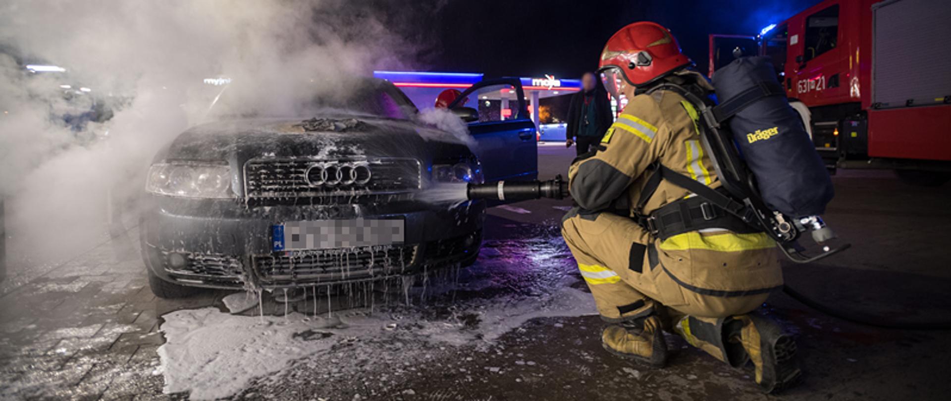 Samochód osobowy marki Audi, spod maski wydobywa się dym. Strażak w aparacie powietrznym kuca przed pojazdem i podaje prąd wody pod maskę.