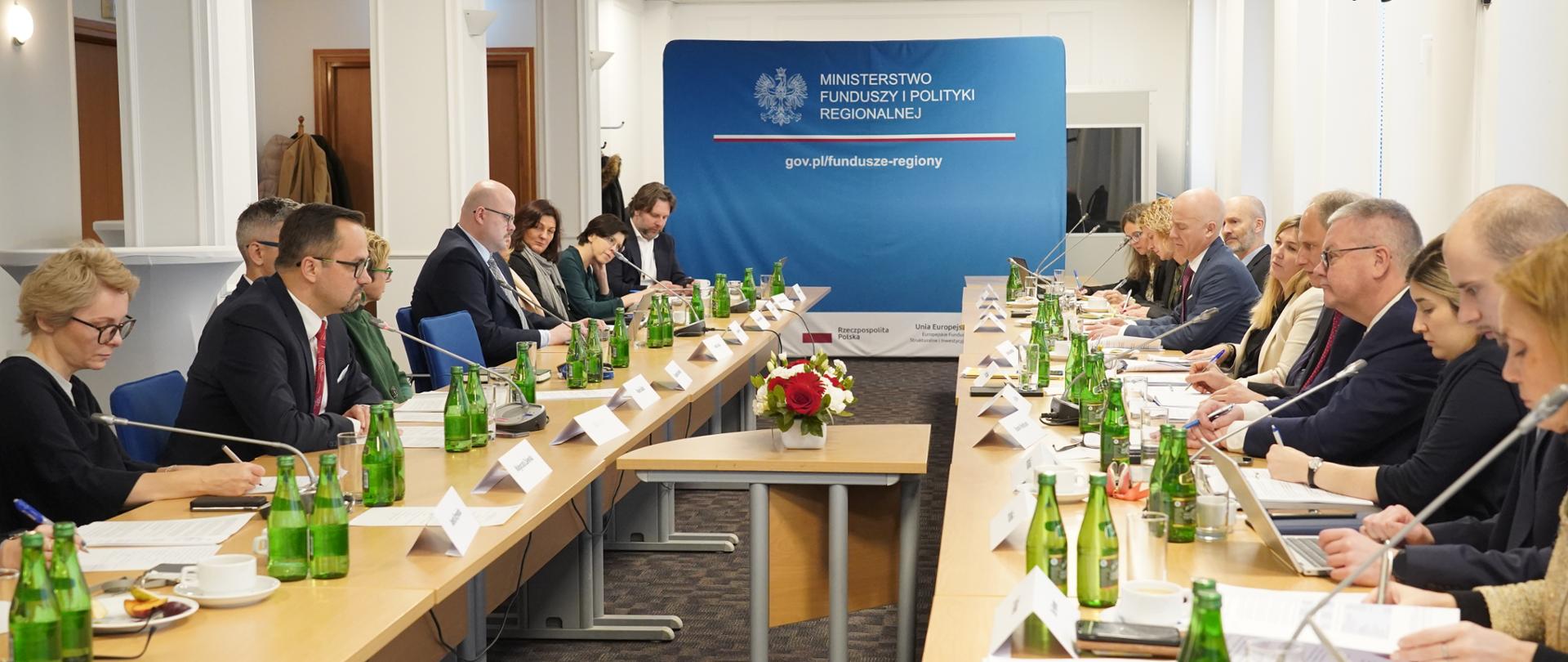 Grupa ludzi siedzi obok siebie przy stole z lewej strony i przy sole z prawej strony.