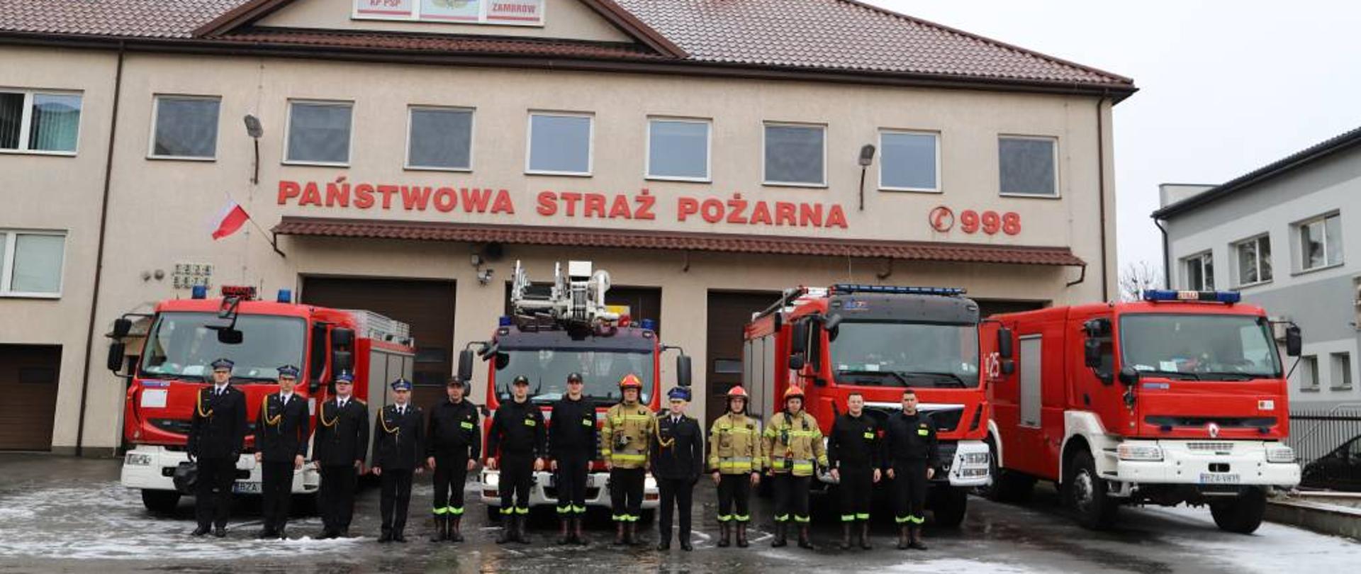 Zdjęcie przedstawia starszego aspiranta Dariusza Hańczuka w mundurze wyjściowym ze sznurem galowym podczas pozowania do pamiątkowego zdjęcia na tle pojazdów będących na wyposażeniu Jednostki Ratowniczo-Gaśniczej w Zambrowie. Obok starszego aspiranta Dariusza Hańczuka z w szeregu stoi zmiana służbowa ubrana w ubrania koszarowe, niektórzy dodatkowo w kurtkę od ubrania specjalnego. Do zdjęcia ustawili się również ubrani w mundury wyjściowe ze sznurem galowym: dowódca Jednostki Ratowniczo-Gaśniczej w Zambrowie starszy Kapitan Paweł Gwardiak oraz poczet sztandarowy. Zdjęcie wykonano w pochmurny dzień na placu Komendy Powiatowej Państwowej Straży Pożarnej w Zambrowie. W tle poza widocznym w części budynkiem komendy widać wystawione z garażu cztery samochody bojowe. 