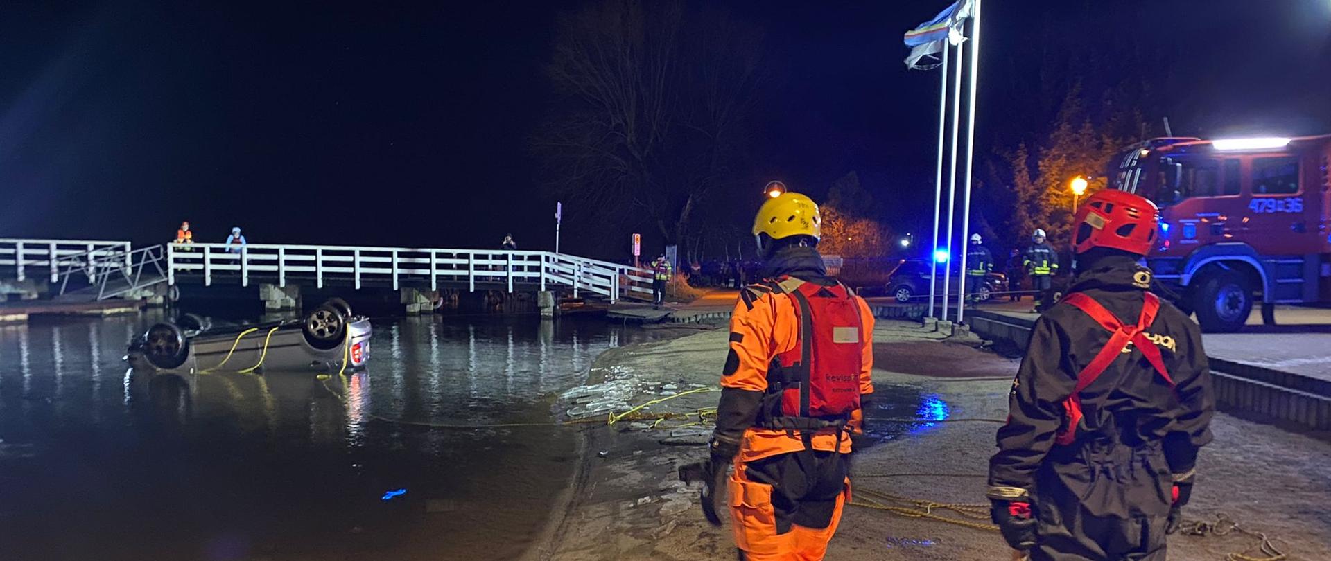 Na zdjęciu widać samochód osobowy leżący na dachu, który wyciągany jest przy pomocy liny. Po prawej stronie kadru na brzegu stoi dwóch ratowników ubranych w skafandry do ratownictwa wodnego. Zdjęcie wykonane nocą.