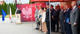 Strażak przemawia przy mównicy na tle czerwonej ścianki z godłem RP pod czerwonym namiotem funkcjonariusze w mundurach oraz osoby w ubraniach cywilnych