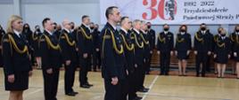 Na zdjęciu funkcjonariusze ubrani w mundury galowe, stojący w szeregu, w tle baner z napisem 30 lecie Państwowej Straży Pożarnej 