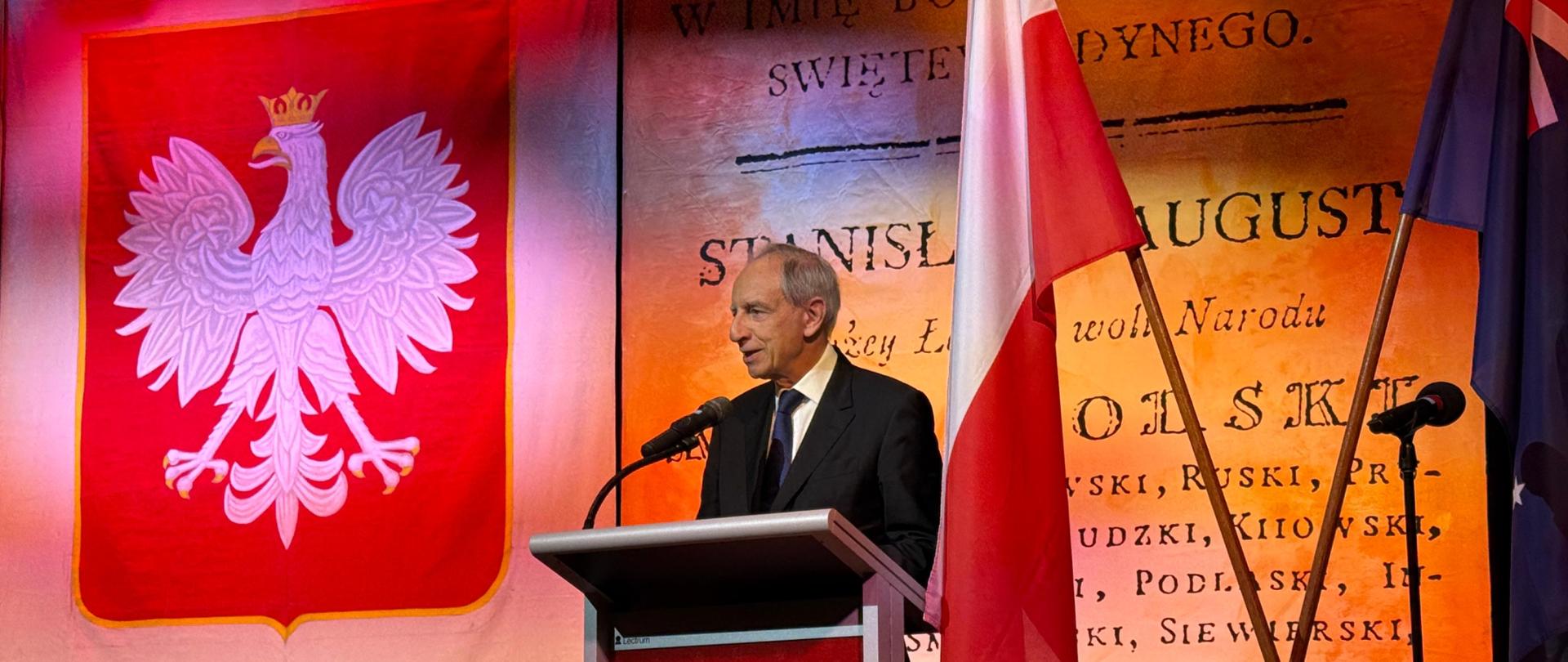Minister przemawia na scenie, w tle godło Polski i flagi Polski oraz Australii.