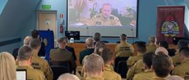 Na sali, w rzędach siedzą oficerowie PSP - ok. 30 osób, w pierwszych rzędach goście narady. Widok na stół prezydialny i ekran, gdzie w ramach transmisji wideo rozmówcą jest zastępca komendanta głównego. Strażacy w mundurach służbowych - musztardowych z czarnym napisem Paśńtwowa Straż Pożarna
