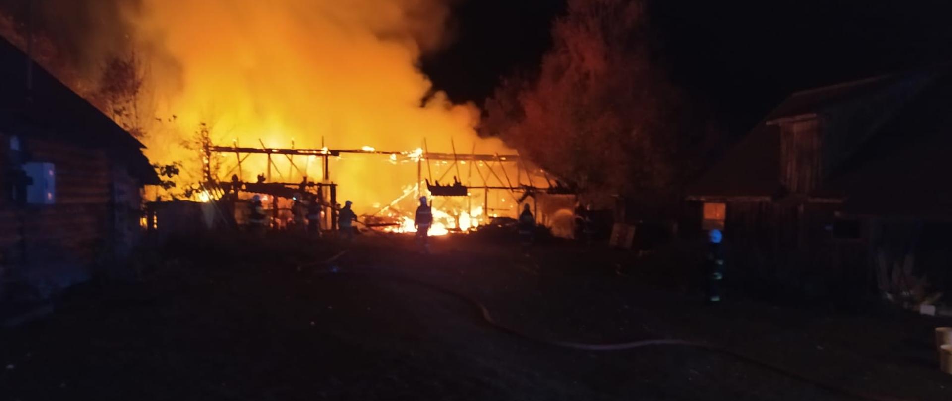Na zdjęciu widać płonąca konstrukcję budynku gospodarczego, płomienie słabną, strażacy gaszą pożar, rozwinięte są dwie linie gaśnicze.