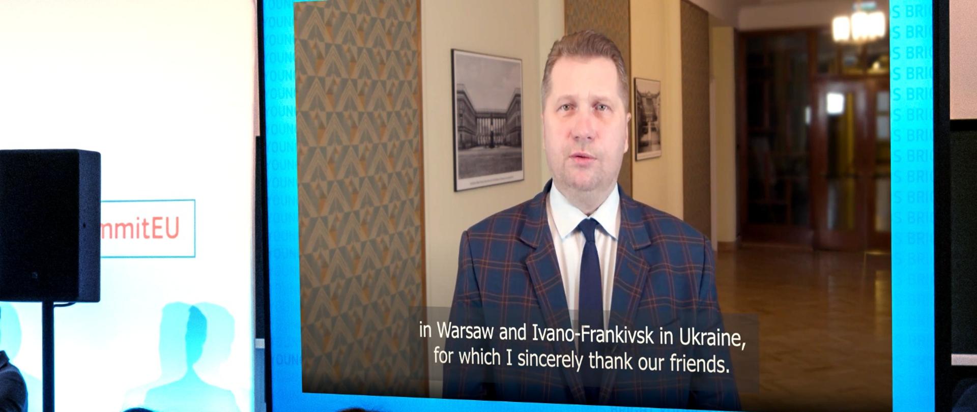 Na sali siedzi wiele osób i patrzy na stojący przed nimi wielki monitor, na którym widać ministra Czarnka na tle korytarza.