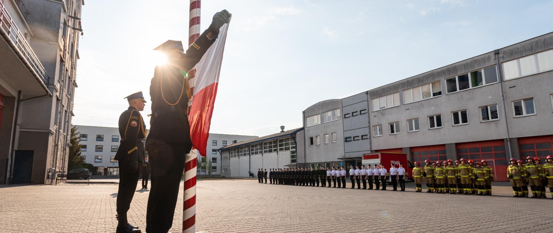 widok na plac szkoły aspirantów, po lewo strażacy dokonują uroczystego wciągnięcia flagi na maszt, po prawo w szeregu stoją strażacy, w tle budynki szkoły