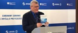 Mężczyzna stoi za szarą mównicą ozdobioną godłem Polski oraz napisem „Główny Inspektorat Sanitarny”. W ręku trzyma zbroszurowany oficjalny raport lub dokument programowy, prezentując go zgromadzonym dziennikarzom.