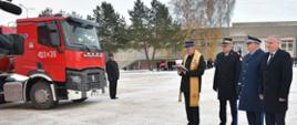 Kapelan strażacki trzyma w ręce modlitewnik i czyta obok stoją wojewoda pomorski pomorski komendant wojewódzki Państwowej Straży Pożarnej oraz komendant wojewódzki Policji w Gdańsku przed nimi stoi samochód specjalny. 