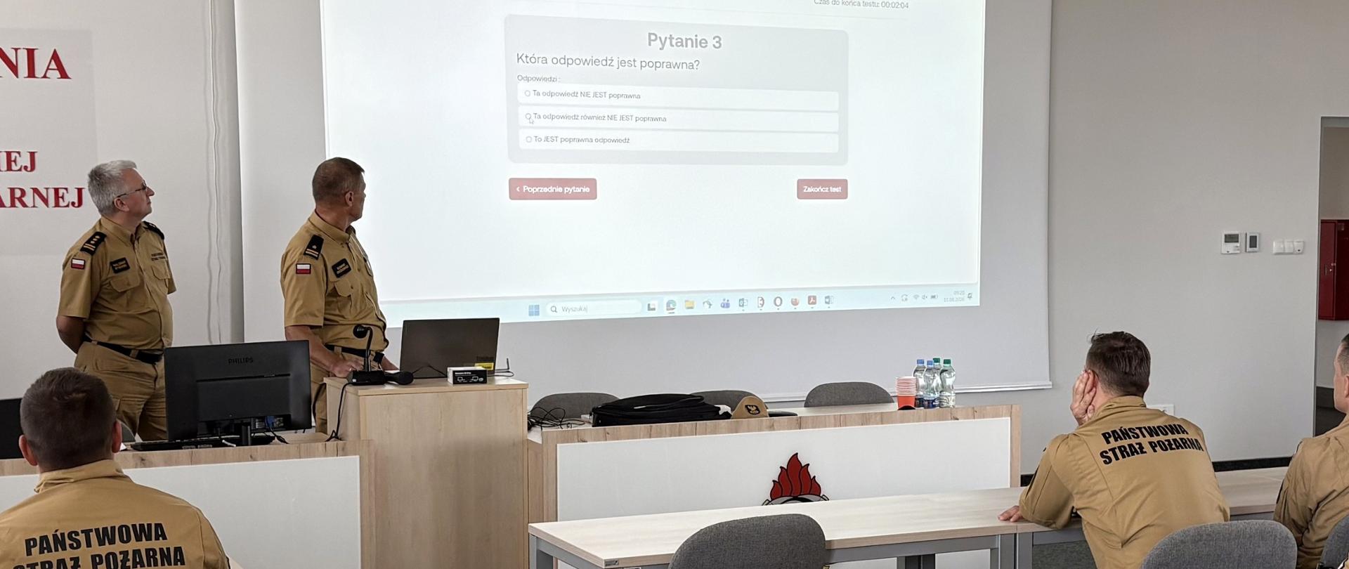 Zdjęcie przedstawia salę szkoleniową podczas prezentacji. Na pierwszym planie, tyłem do obiektywu, siedzą dwaj strażacy w beżowych mundurach z widocznym napisem na plecach „PAŃSTWO STRAŻ POŻARNA” oraz krzesła i ławki szkolne. Z przodu sali, obok jasnej mównicy i ekranu projekcyjnego, stoją dwaj mężczyźni w mundurach służbowych – jeden z nich obsługuje laptopa i patrzy na ekran, na którym wyświetla się test z pytaniem i odpowiedziami do wyboru. W tle widoczny jest jasny, minimalistyczny wystrój sali z wykładziną podłogową.