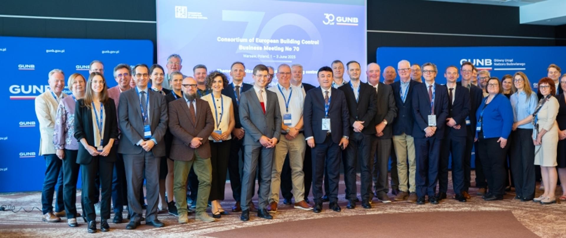 Grupa kilkudziesięciu uczestników pozuje do wspólnego zdjęcia podczas 70. spotkania Konsorcjum Europejskiego Nadzoru Budowlanego w Warszawie, organizowanego przez Główny Urząd Nadzoru Budowlanego (GUNB). Uczestnicy stoją na tle niebieskich roll-upów i ekranu z nazwą wydarzenia. Osoby ubrane są w stroje biznesowe, wiele z nich ma identyfikatory konferencyjne na smyczach.