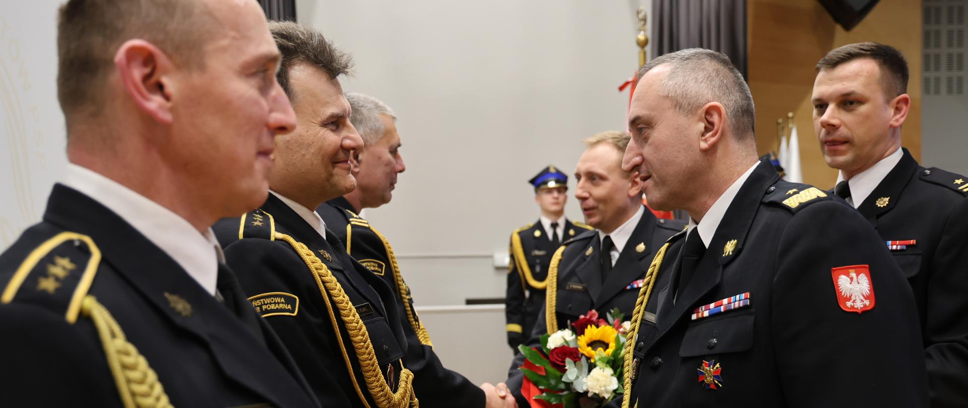 Bliskie ujęcie momentu składania gratulacji. Funkcjonariusz w mundurze wyjściowym wymienia mocny uścisk dłoni z jednym z odchodzących na emeryturę strażaków. W tle widoczny jest inny funkcjonariusz trzymający kolorowy bukiet kwiatów. Na pierwszym planie wyraźnie widać złote pagony i detale umundurowania galowego.