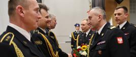 Bliskie ujęcie momentu składania gratulacji. Funkcjonariusz w mundurze wyjściowym wymienia mocny uścisk dłoni z jednym z odchodzących na emeryturę strażaków. W tle widoczny jest inny funkcjonariusz trzymający kolorowy bukiet kwiatów. Na pierwszym planie wyraźnie widać złote pagony i detale umundurowania galowego.