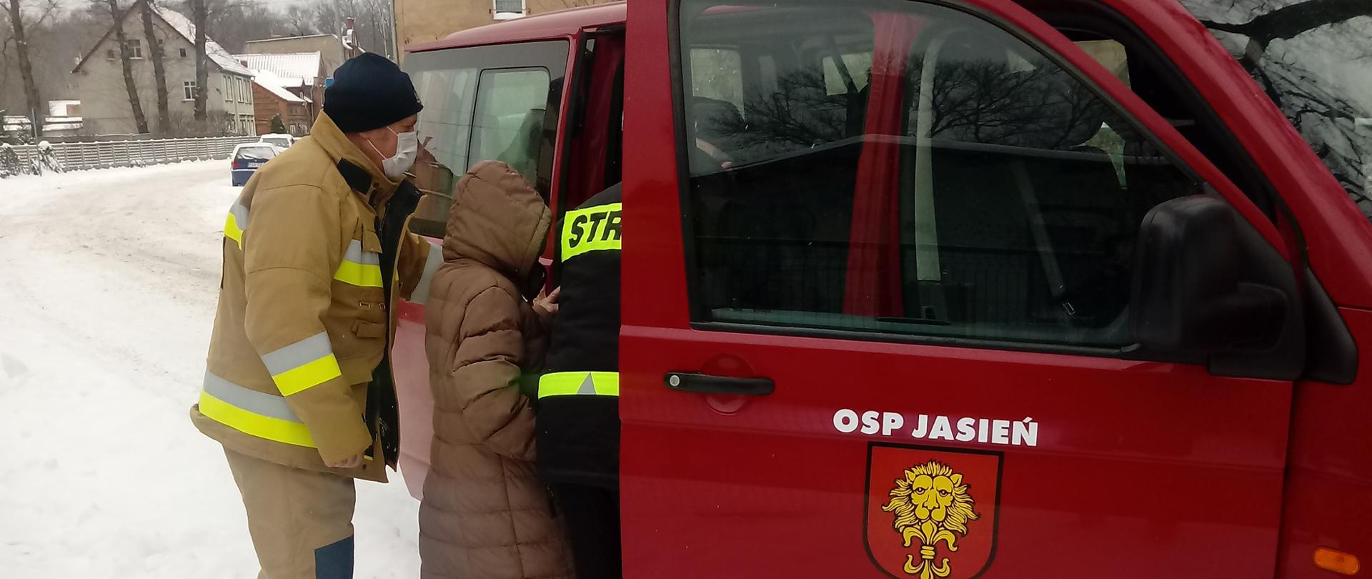 Warunki zimowe, gruba warstwa śniegu zakrywa chodniki i drogę. Do czerwonego samochodu typu bus wsiadają seniorzy którzy będą zawiezieni przez strażaków na szczepienia przeciwko Covid dziewiętnaście. Przy czerwonym busie dwóch strażaków pomaga wsiąść seniorowi. 