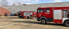 w tle hala produkcyjna, przed nią stoją dwa pojazdy pożarnicze, widać rozwinięte węże