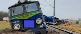 Zdjęcie przedstawia lokomotywę wykolejoną z toru oraz kilka jej wagonów także wykolejonych. Obok lokomotywy znajdują się przedstawiciele różnych służb i instytucji prowadzący działania. 