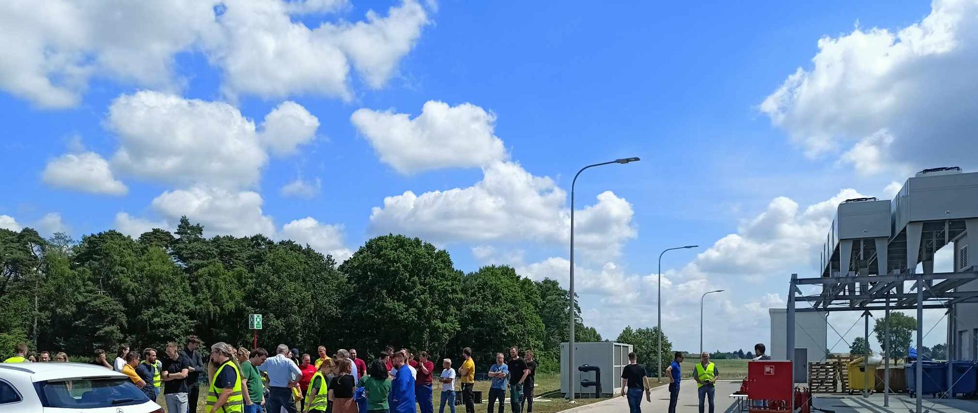 Pracownicy hali objętej działaniami zbierają się w miejscu wyznaczonym do zbiórki podczas ewakuacji obiektu. Trwa liczenie pracownikow. 